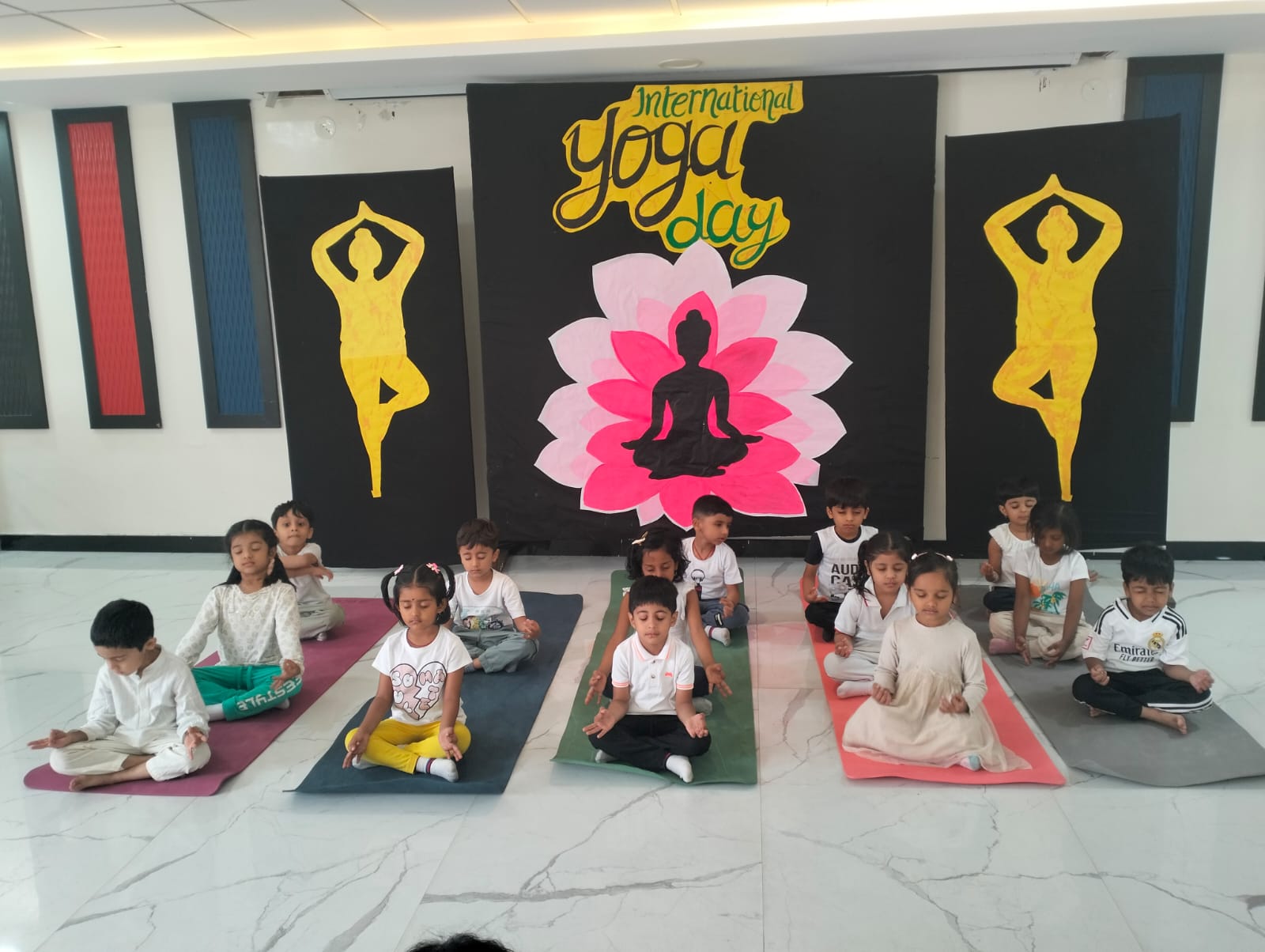 Primary students celebrating International Yoga Day, performing a meditation pose on mats in front of a themed backdrop.