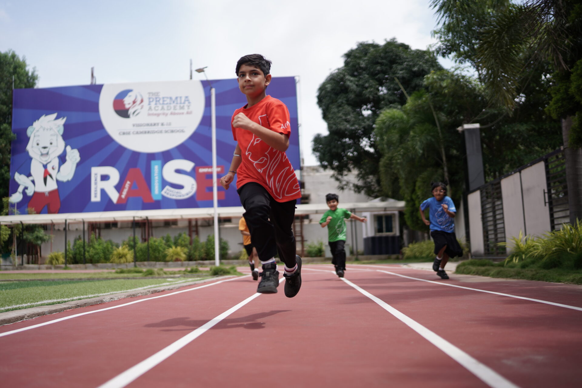 Premia Academy primary students sprinting on the professional outdoor running track with the 'RAISE' billboard visible in the background.