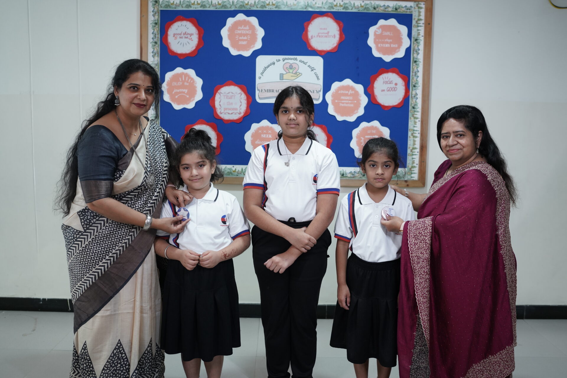 Two female teachers pin badges on two primary students with another student standing between them in front of a bulletin board