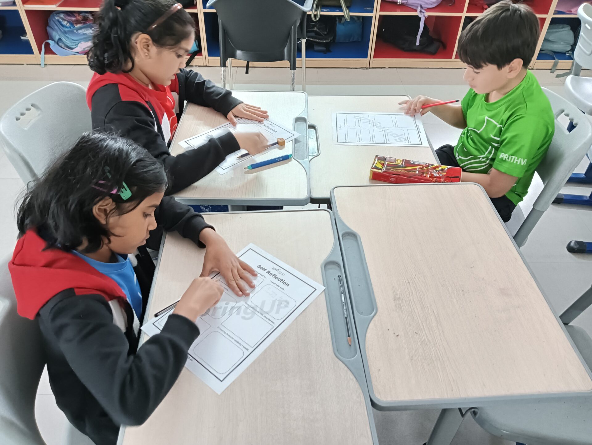 Three young students sitting at desks in a classroom, focused on completing worksheets