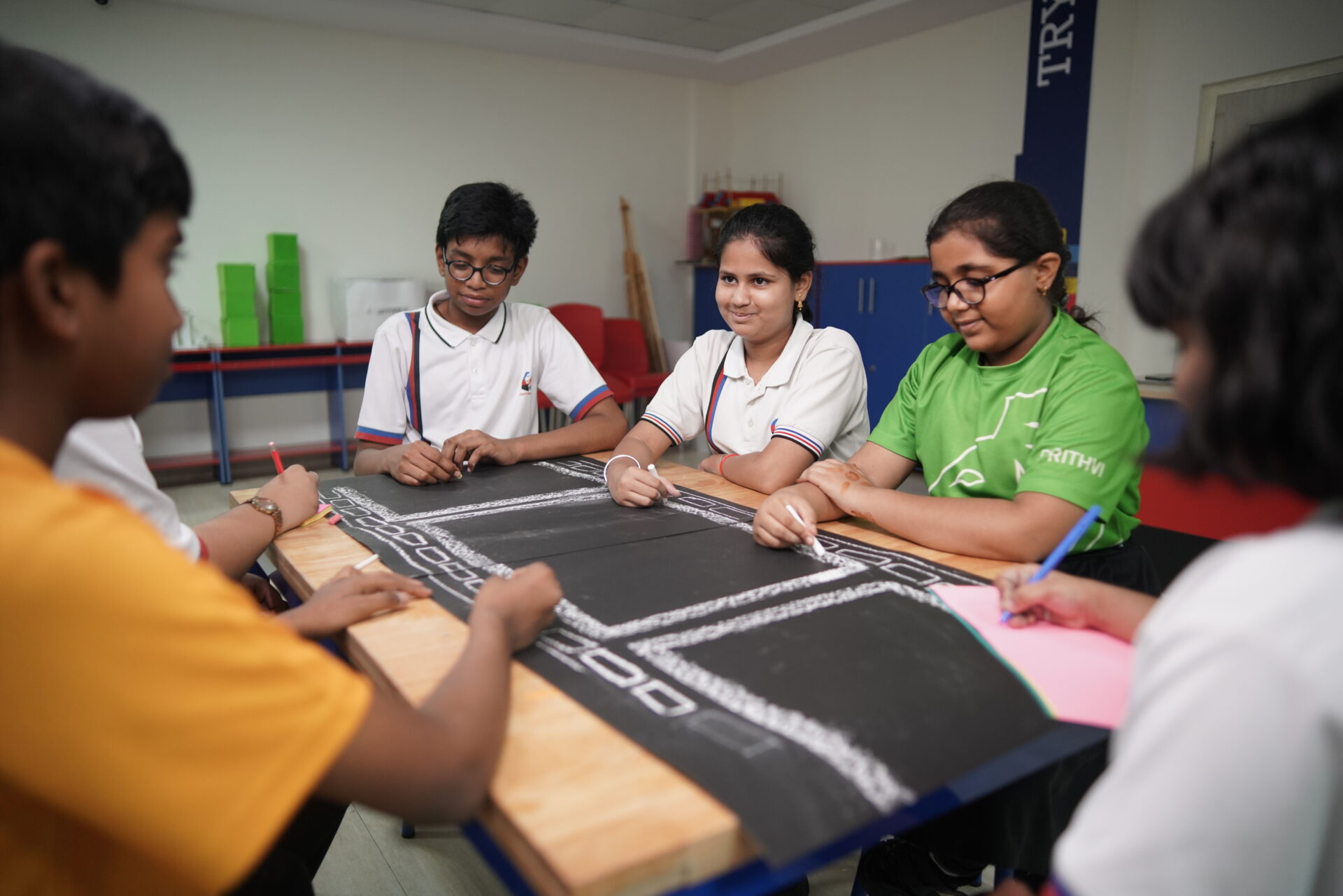 Premia Academy students collaborating on a group project, drawing a road or circuit design onto black paper in the classroom.