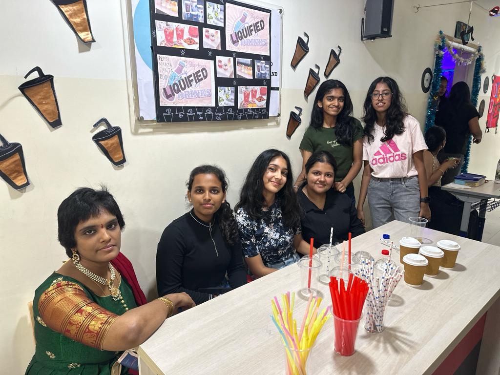 Teacher and five female secondary students at a table with cups, straws, and a 'Liquified Drinks' display board