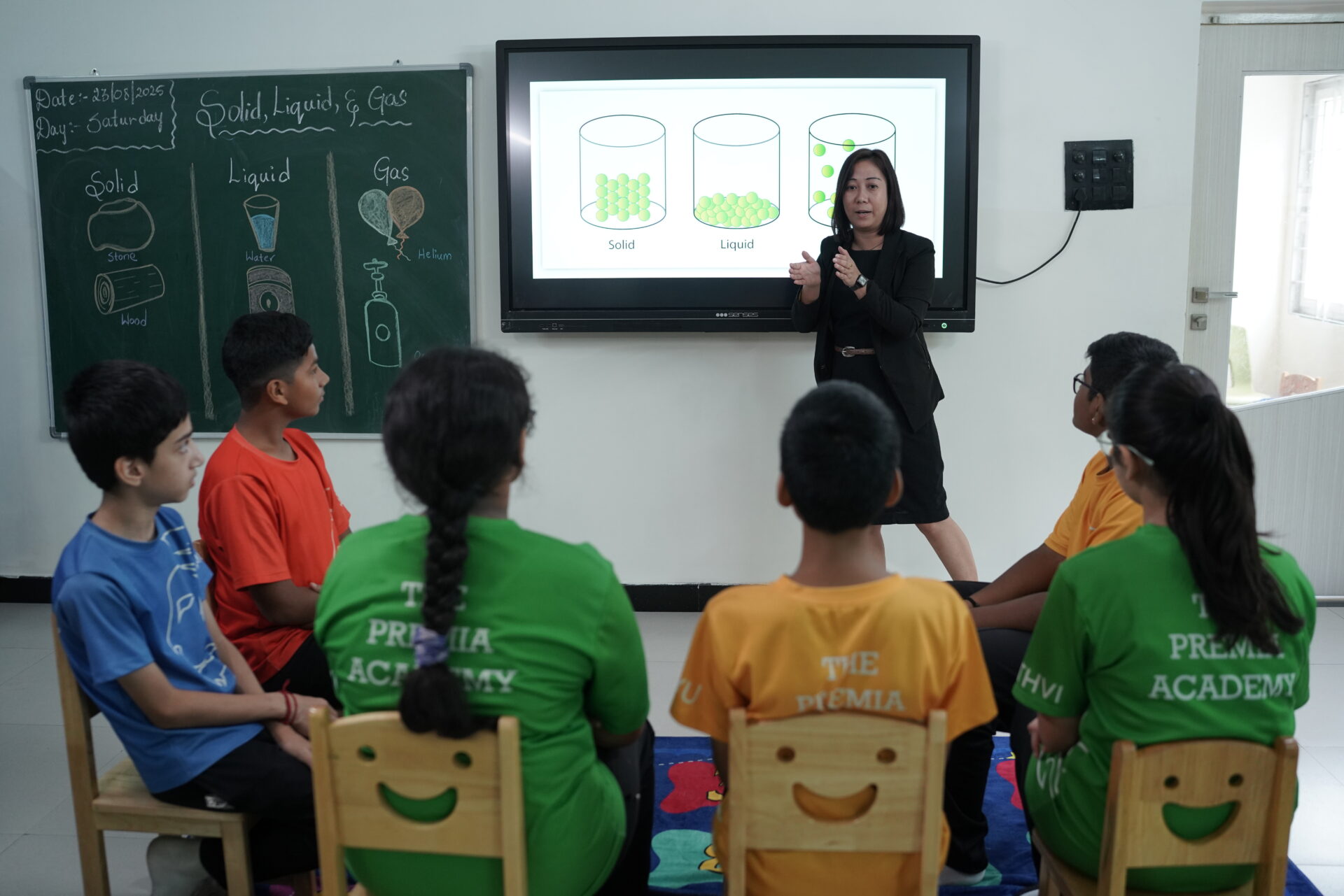 Teacher using a digital screen and chalkboard visuals to teach middle school students about the molecular arrangement of Solid, Liquid, and Gas.