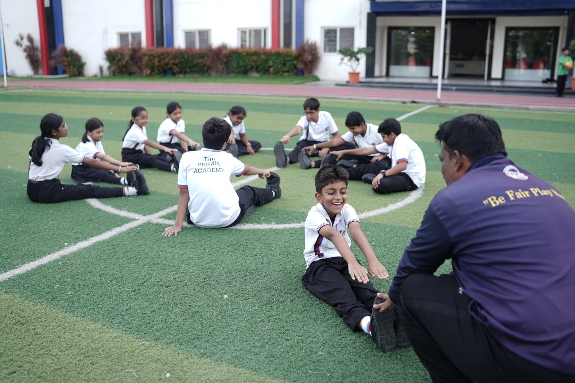 Premia Academy students performing warm-up stretching exercises on the artificial turf sports field, supervised by a coach.