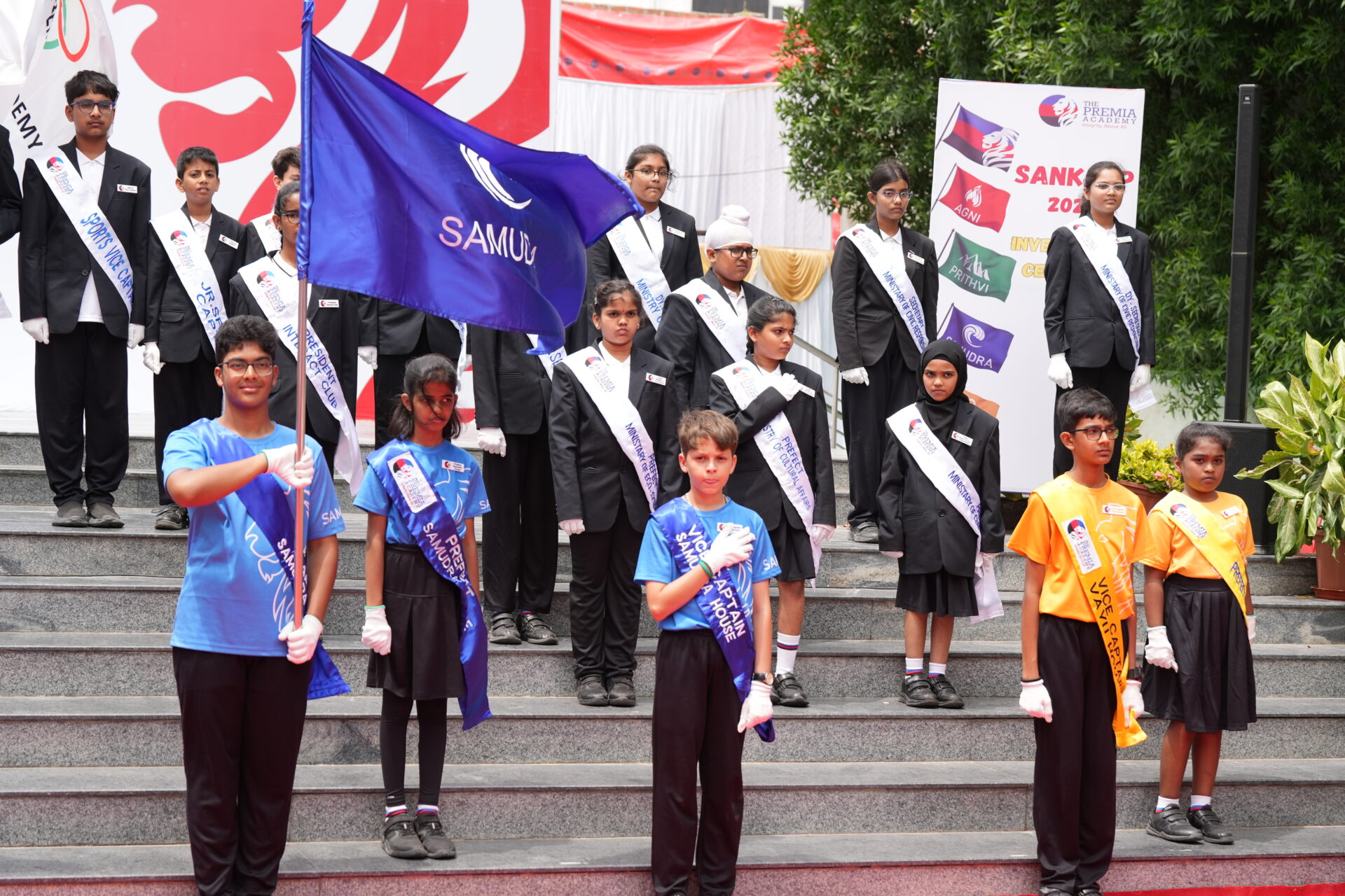 Students in uniform and sashes holding flags and standing on steps for a school event ceremony