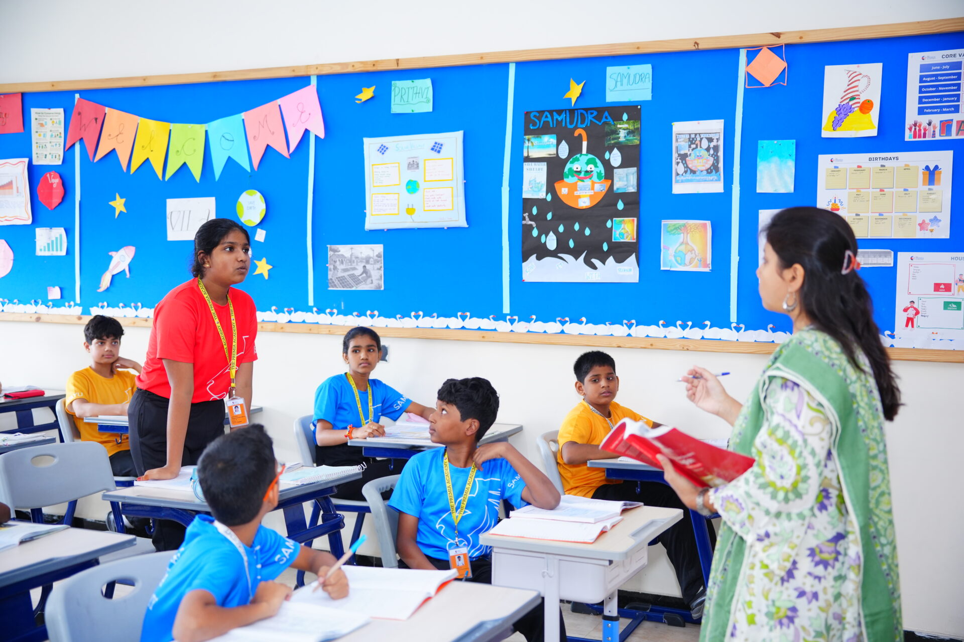 Premia Academy middle school students receiving team-teaching instruction in a classroom featuring house system (Samudra, Prithvi) display boards.