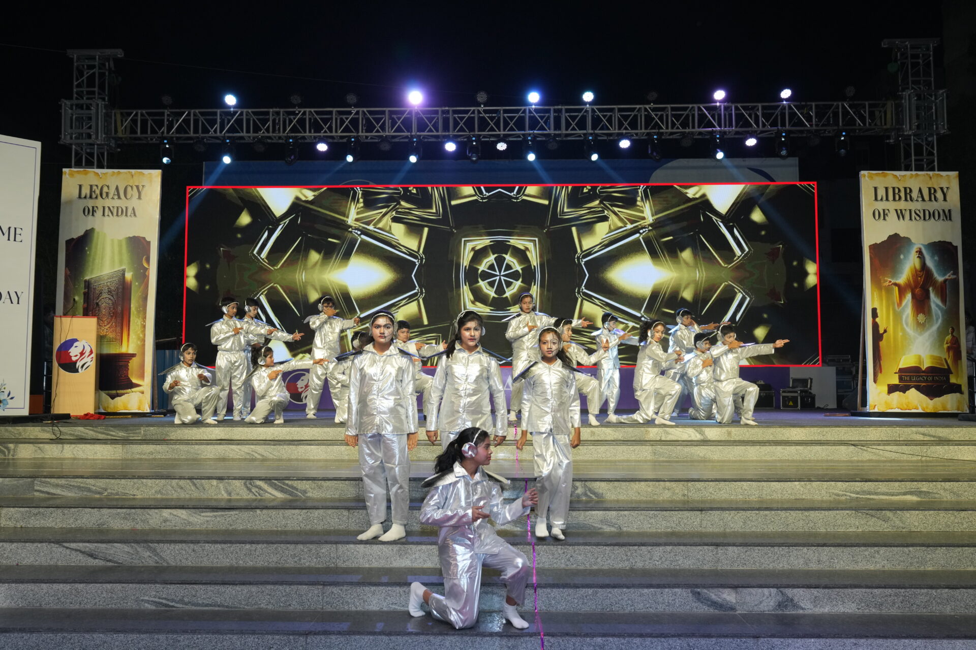 Students in futuristic silver costumes perform a choreographed dance routine on an outdoor stage at night