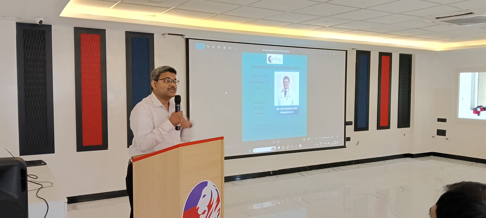 speaker addresses an audience from a podium with a screen displaying a presentation about 'Growing Up Gracefully'