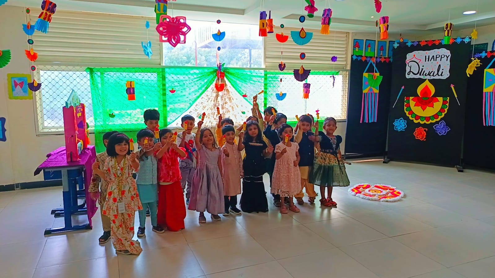 Primary students celebrating Diwali in the classroom, holding decorative lights and standing near a rangoli